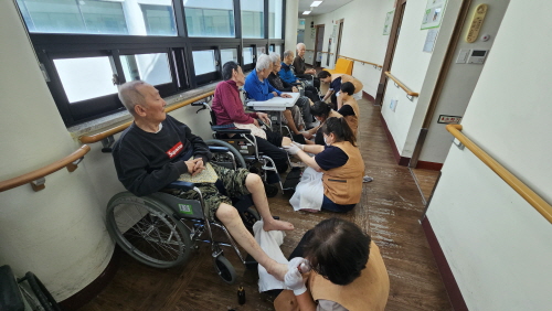 남자 어르신들 5명이 휠체어에 앉아 계시고 발마사지를 받고 있다.
