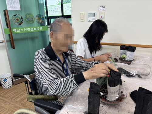 남자 어르신이 숯을 두손을 만지고 있고, 그 뒤로 여학생이 보인다.