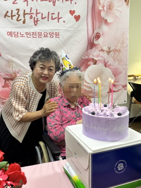 꼬깔모자를 쓴 여자 어르신과 다른 한분의 여자 어르신이 생일케이크를 앞에 두고 사진 찍는 모습