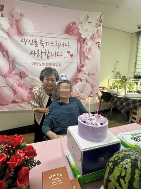 꼬깔모자를 쓴 남자 어르신과 여자 어르신이 생일케이크를 앞에 두고 사진 찍는 모습