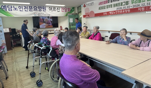 정면에는 현수막이 걸려있고, 화면에는 영상이 재생되고, 20여명이 사람들이 테이블에 앉아있고, 2명이 서있다