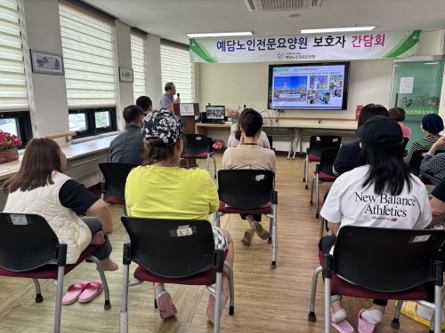정면에 현수막과 TV 화면이 보이고 10여명이 앉아 있으며 여자분이 화면을 가르키며 설명하고 있다