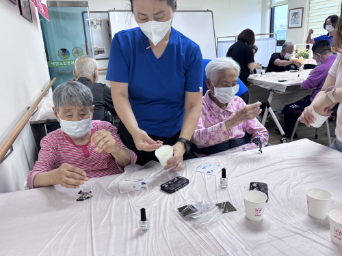마스크를 쓴 여자 어르신 두분이 테이블 위에서 무언가를 만들고 옆에서 여자분이 도와주고 있다.