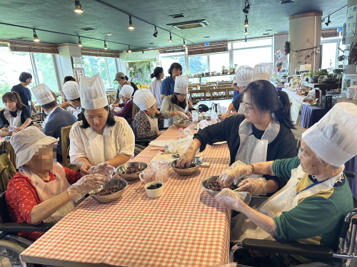 20여명의 사람들이 요리사 모자를 쓰고 비닐장갑을 끼고 볼에 담긴 블루베리를 섞고 있다.