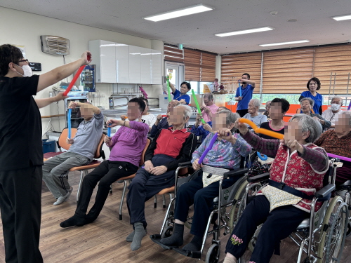 여자 강사분이 두 손으로 튜브를 잡고, 10여명의 어르신들이 동작을 따라하고 있다.