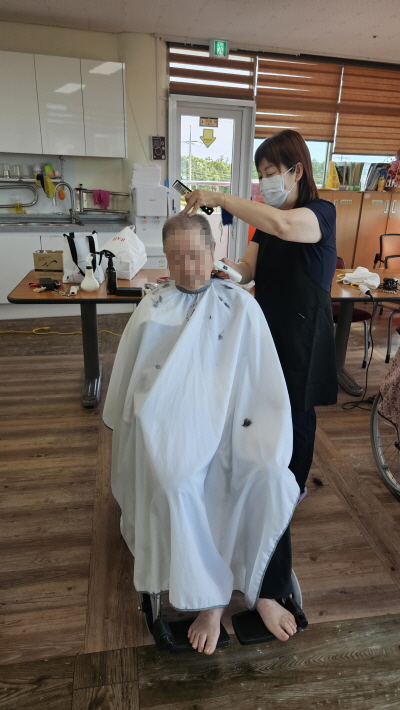 가운을 입은 어르신이 휠체어에 앉아 계시고 미용사분이 어르신 머리를 다듬어주고 있다