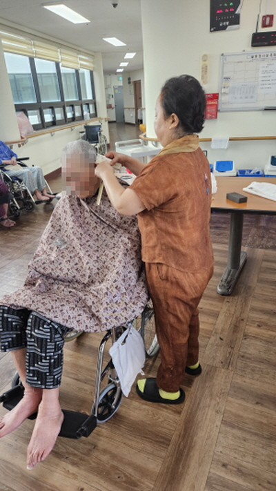 가운을 입은 어르신이 휠체어에 앉아 계시고 미용사분이 어르신 머리를 다듬어주고 있다